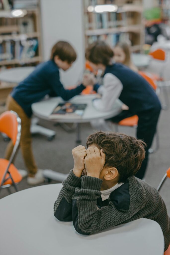 A disheartened child sits alone in a school library while other kids interact.